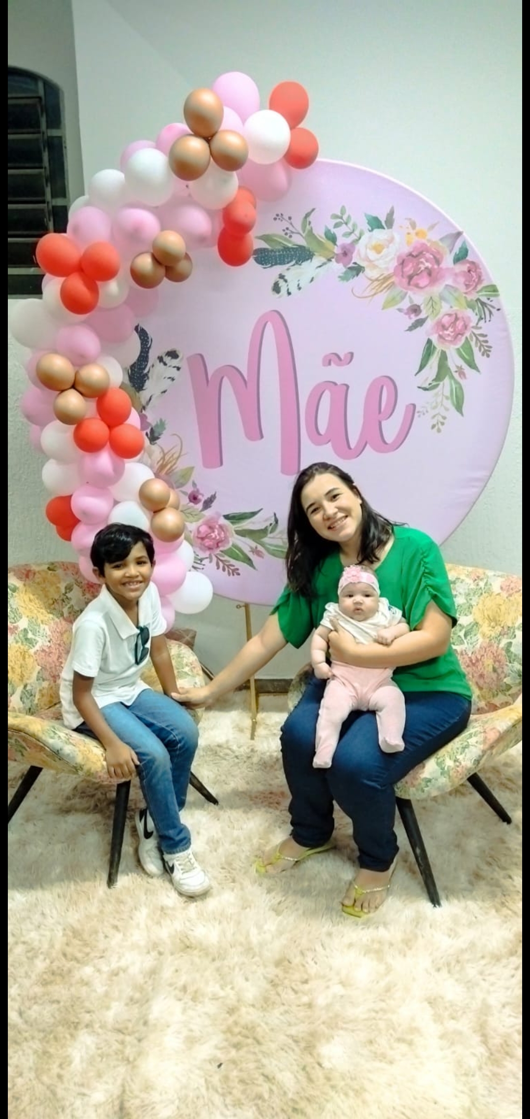 Jéssica sentada com os filhos e um painel atrás escrito Mãe